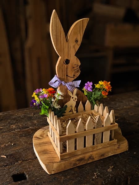 Stehender Osterhase aus Holz in einem kleinen Zaunbeet mit Blumen und Heu
